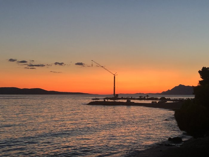 Solnedgång över Medelhavet med silhuett av en byggkran och berg i bakgrunden.