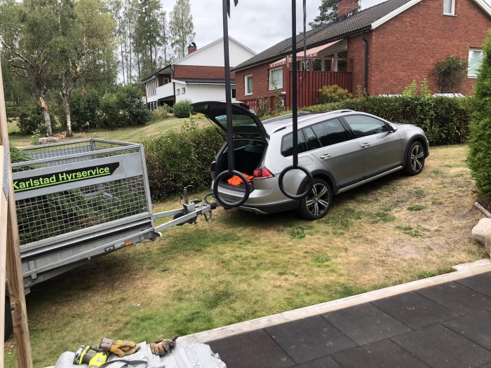 Bil parkerad vid tomtgräns bredvid en borttagen buske, öppet bagagelucka med släpvagn, bakgrunden visar en hammock och hus.