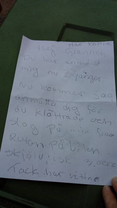 Handskriven lapp med meddelande om skadad bil och vittnesuppgifter, hållen ovanpå en grön yta.