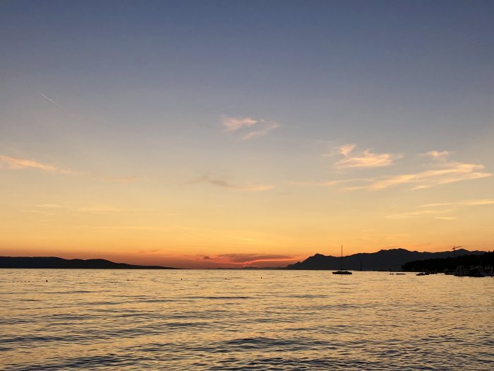 Solnedgång över havet med bergssilhuetter och en segelbåt mot en himmel med varma färger