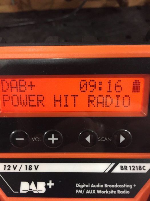 Närbild på en orange DAB+ arbetsplatsradio som visar POWER HIT RADIO på displayen och knappar för volym och kanalsökning.