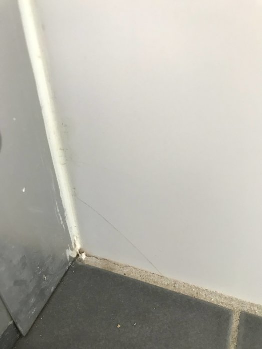 Spricka i vit kakelplatta nära golv och bristfällig fogning i duschhörna med gråa plattor.