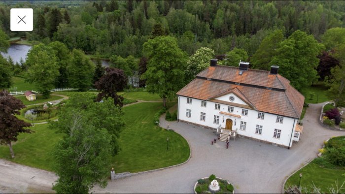 Flygbild på en stor vit herrgårdsbyggnad omgiven av gröna trädgårdar och skog nära vatten.