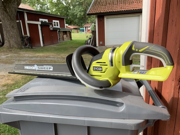 Gul och grå batteridriven häcksax från Ryobi placerad på en grå soptunna framför en röd stuga och garage.