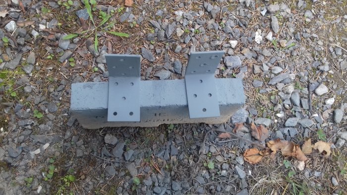 Två grå målade vinkelbeslag monterade på en betongtrallbit som ligger på grusig mark.