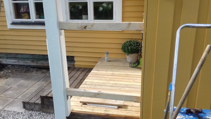 Nytt trätrall med oljad kant och påbörjat räckesbygge vid sidan av en gul husvägg med detaljer och verktyg synliga.