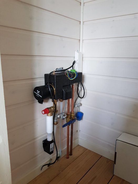 Nyinstallerat golvvärmesystem med kopplingar och regulator i ett hörn av ett rum med träbeklädnad och -golv.