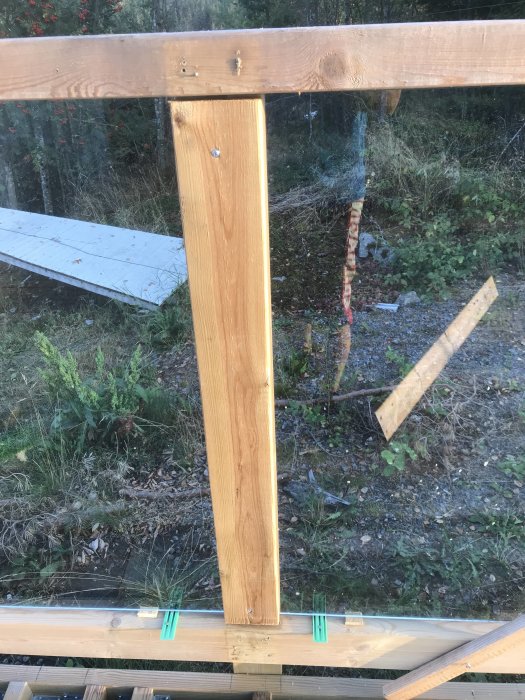 En träregel fastklämd mellan två glasskivor i ett räcke, mot en bakgrund av grönska och byggmaterial.