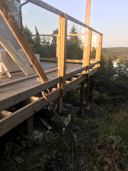 Heltäckande bild av ett trädeck med ett räcke under konstruktion och glaspaneler vid solnedgången med skog och en sjö i bakgrunden.