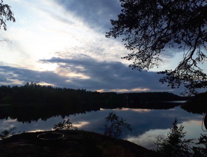 Solnedgång över en sjö i Sörmland med spegling i vattnet, omgiven av träd och moln.