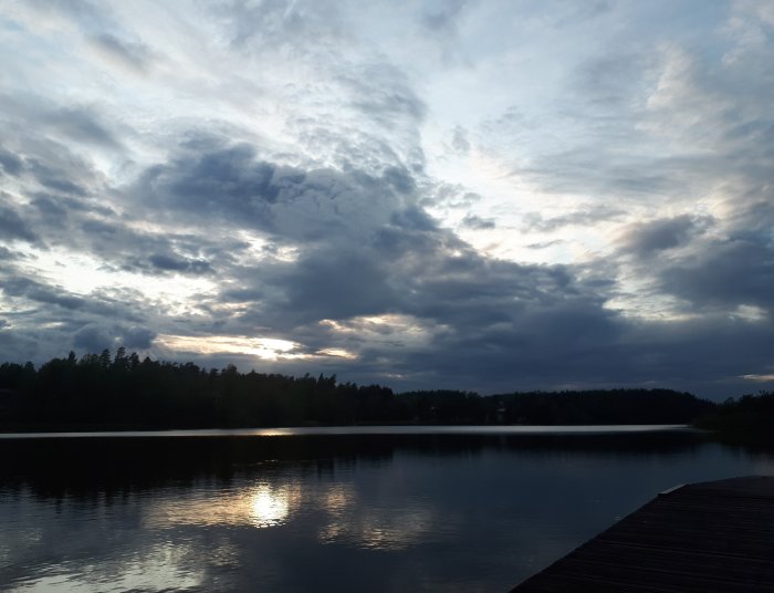 Solnedgång över en lugn sjö i Sörmland med dramatisk molnhimmel och träbrygga i förgrunden.