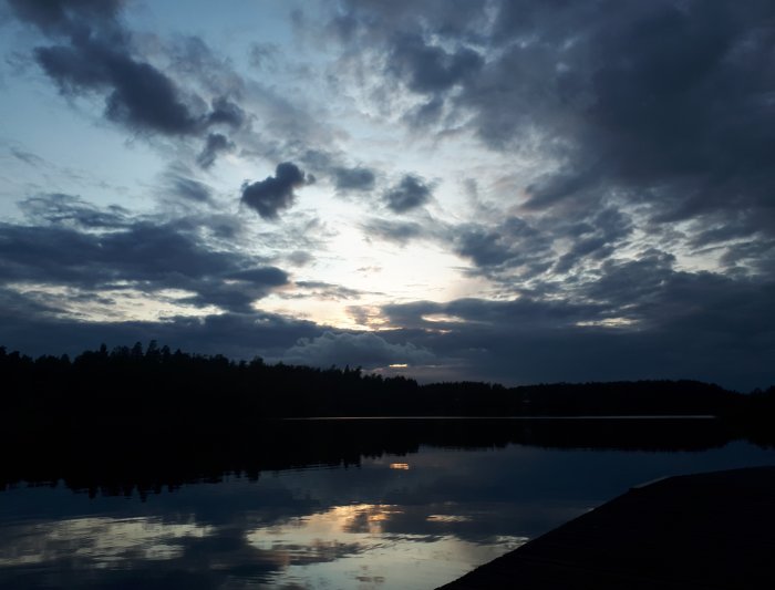 Solnedgång med dramatisk himmel reflekterad i lugnt vatten vid en sjö i Sörmland, vy från en brygga.