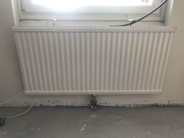 Äldre ribbat element under ett fönster i ett rum med vit vägg och grått golv.