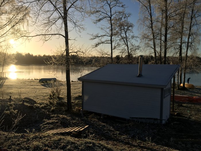 Vinterbild av ett småhus med plåttak och skorsten vid sjökanten i morgonsol.