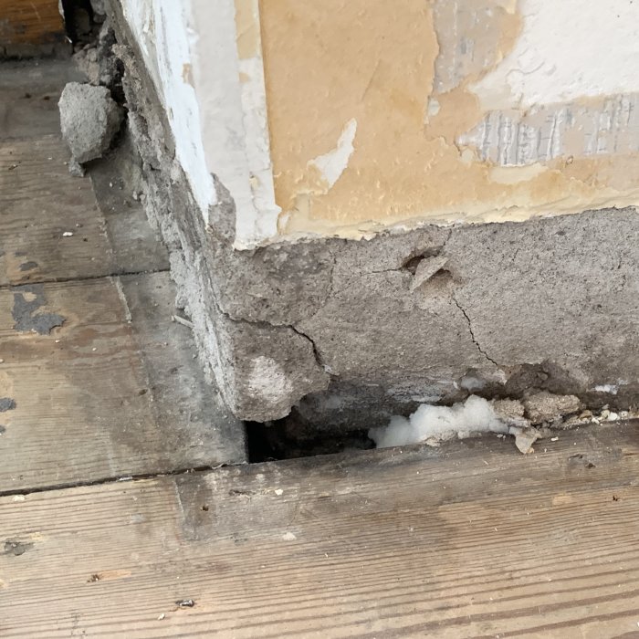 Skadad vägg och golv med borttagen golvsockel där puts har lossnat och lämnat ett hål vid vägghörnan.