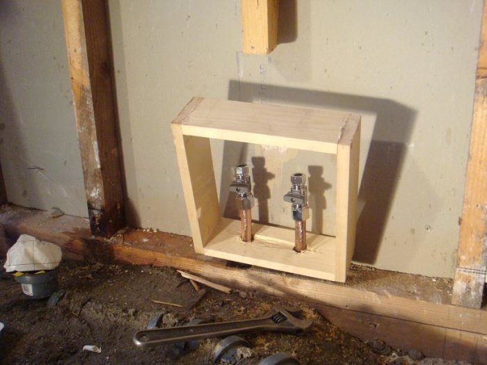 Träram inspektionslucka inbyggd i vägg med rörkopplingar, verktyg på betonggolv.