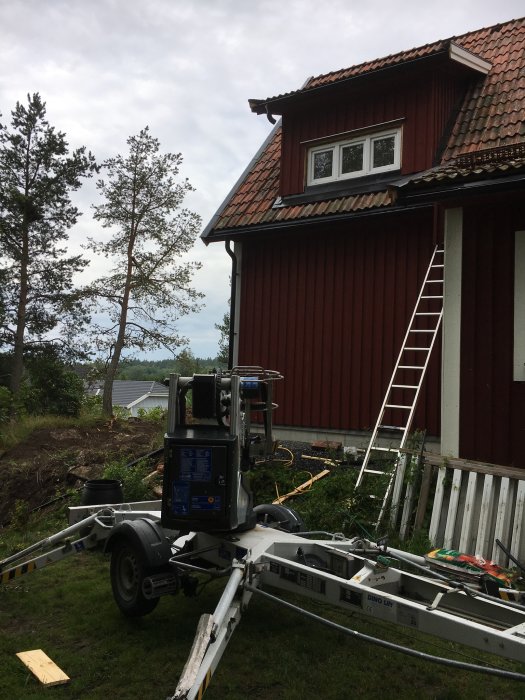 Skylift utrustad för arbete framför ett rödmålat hus med takkupa, omgivet av grönska och en stege lutad mot husväggen.