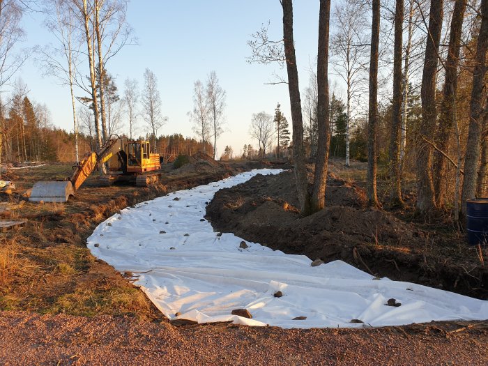 Byggplats med grävmaskin och nylagt fiberduk i en böjd infart genom skogslig mark.