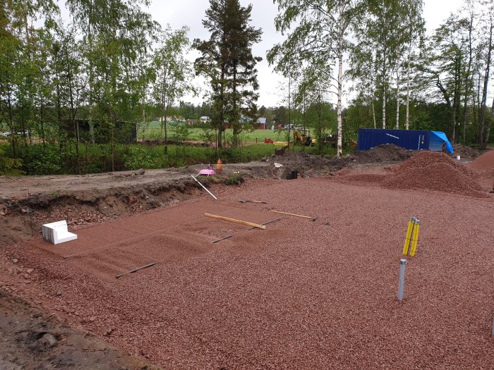 Upparbetad bygggrund med rödaktigt krossgrus, verktyg utspridda och en blå container i bakgrunden.