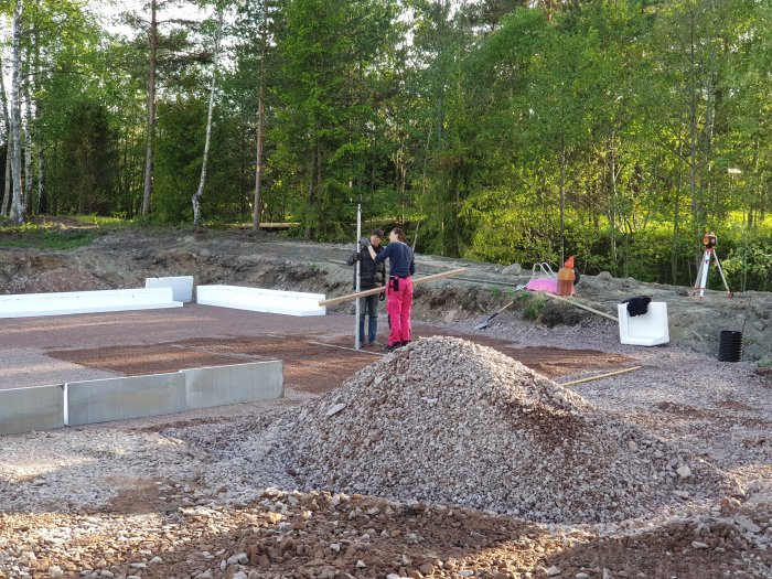 Byggarbetsplats med grushög, kantelement och personer som mäter med rotationslaser.