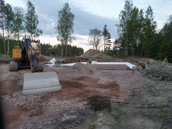 Grävmaskin vid byggarbetsplats med schaktat område och upplagda kantelement för grund.