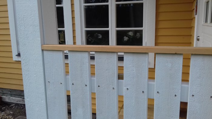 Nybyggt vitmålat trästaket med en överliggare framför en gul husvägg och ett fönster med vita karmar.