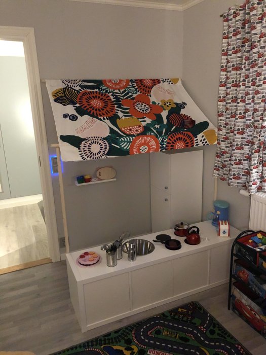Barnlekrum med leksakskök och färgglad panel på väggen, bredvid gardiner med bilar.
