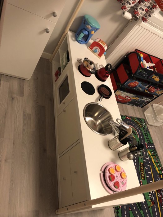 Ett barns lekrum med leksakskök och diverse köksleksaker utspridda, bredvid en bil-matta.