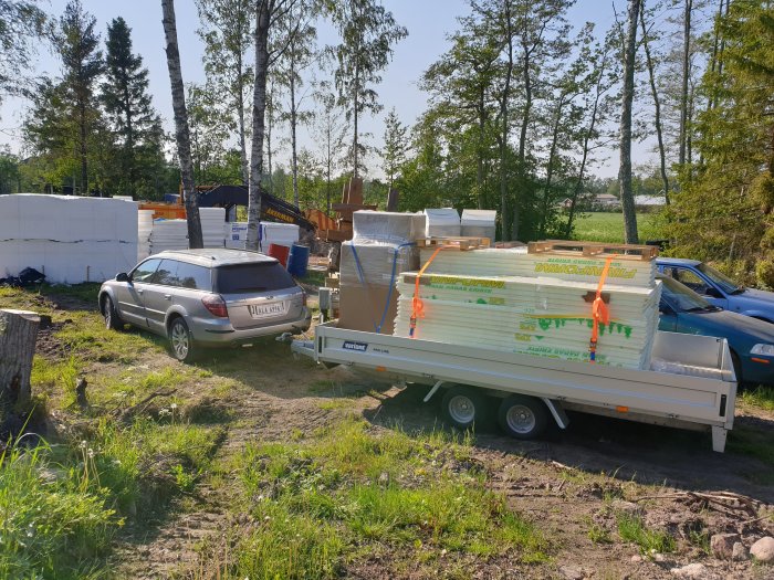 Byggarbetsplats med bil, trailer lastad med isoleringsmaterial och byggmaterial i bakgrunden.