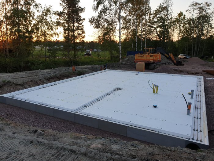 Garageplatta under konstruktion med isolering och armeringsnät, omgiven av grävmaskin och grushögar vid skymning.