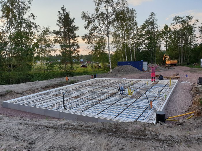 Armeringsnät på garagegrund med isolering och armeringsjärn, i en utgrävd tomt med grävmaskin i bakgrunden.