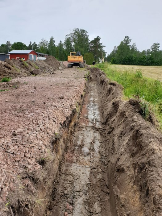 Långt vattengrävt dike med stående vatten och en grävmaskin i bakgrunden, omgiven av högar med grus.
