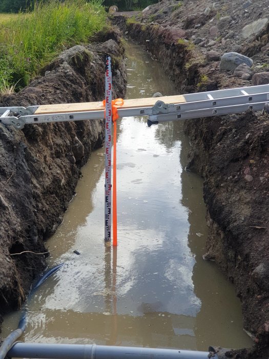 Vattenfyllt dike för VA-anläggning med mätsticka och rörledningar vid byggprojekt.