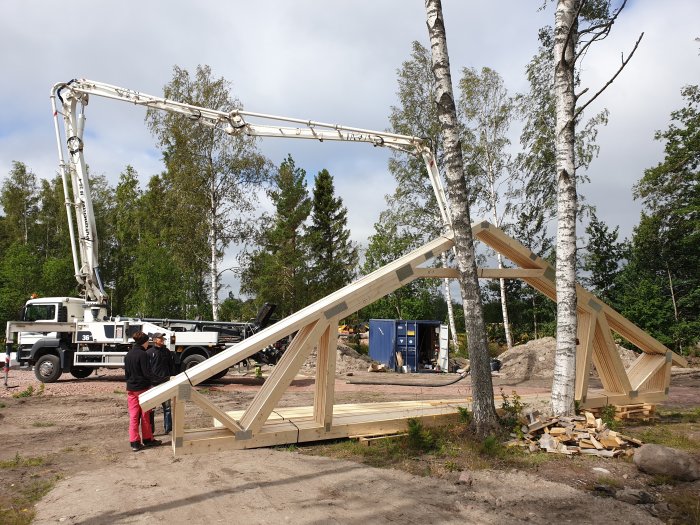 Takstolar till ett garage hålls på plats av en lastbilsmonterad kran medan två personer i arbetskläder diskuterar konstruktionen på en byggarbetsplats.