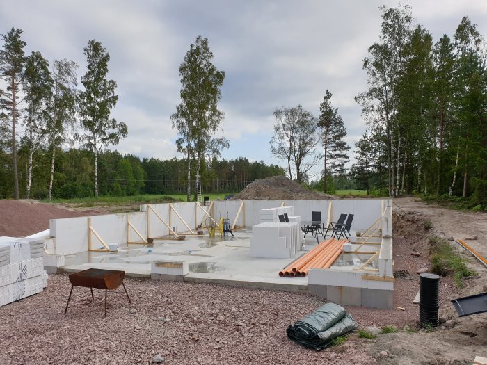 Byggarbetsplats i tidigt skede med betongfundament, väggblock, armeringsjärn och byggmaterial omgivet av träd.