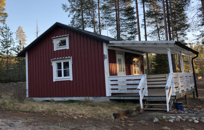 Röd stuga med vit veranda och plasttak vid skogsbryn under tidig solnedgång.