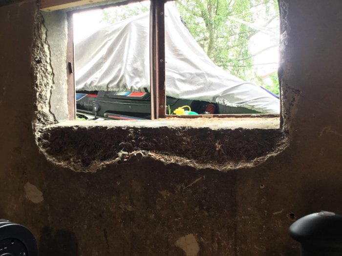 Skadad vägg runt ett fönster som behöver reparation, synliga sprickor och ojämnheter i putsen.