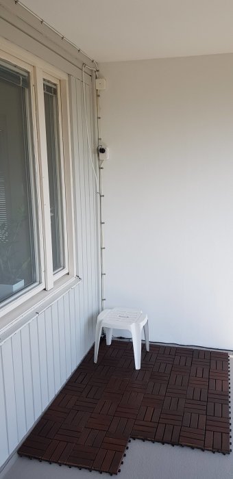Klamring av kablar längs en vit vägg nära fönster ovanför mörkbruna plastplattor på uteplatsen.