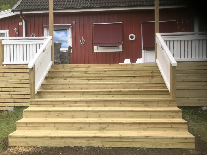 Nybyggda trätrappor som leder upp till en entré med vit träräcke och rött hus i bakgrunden.