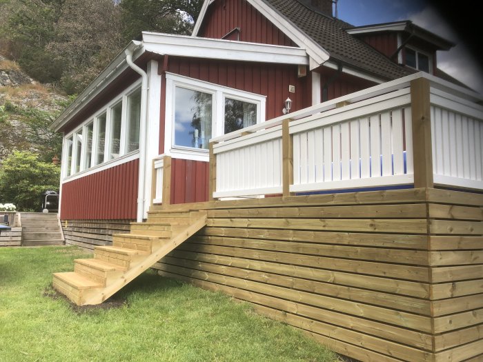 Nybyggd träveranda med vit räcke vid ett rött hus med vita fönster, trappa ner till grönt gräs.