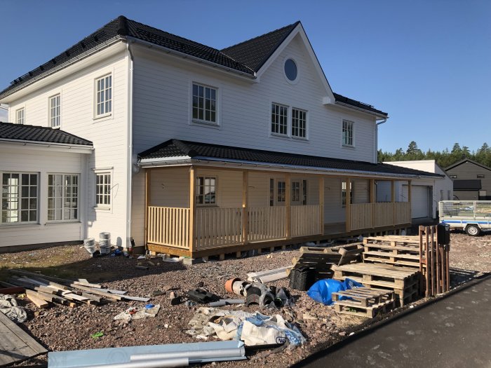 Vitt hus med svart tak och nybyggd träveranda, byggmaterial syns spridda på marken framför huset.