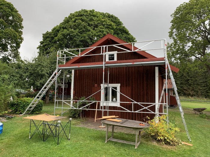 Rött trätorp med byggnadsställning och stege, på en gräsmatta med omgivande träd.