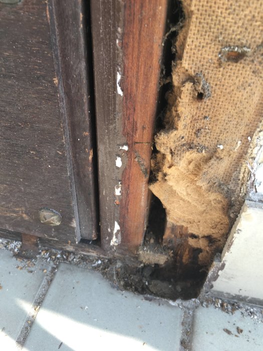 Skadad masonit och fuktig papp bakom trädetaljer vid klinkergolv under veranda