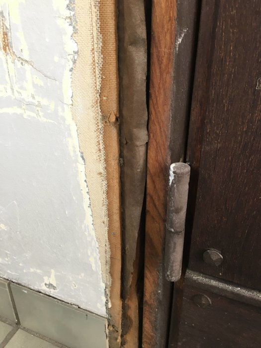 Skadad masonitvägg med blåsning vid golvet och mörk isolering eller papp synlig bakom, intill klinkers.