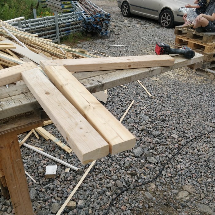 Trätimmer under konstruktion på en grusig yta med verktyg och byggmaterial runtomkring.
