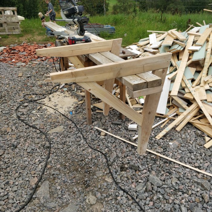 Nybyggt skärmtak av spillvirke vid sågbänk, med virkeshög och verktyg i bakgrunden.
