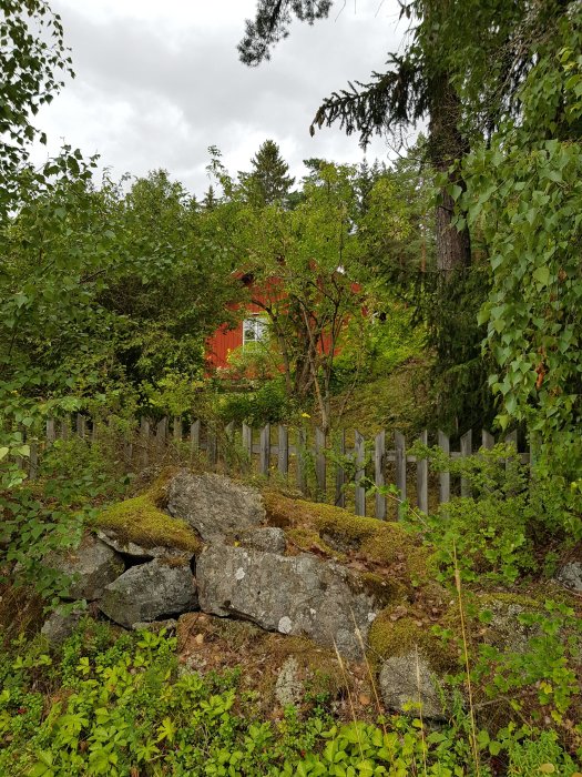 Vy över klippor och grönska med röd stuga i bakgrunden och staket i förgrunden.