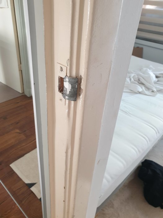 Dörrkarm med urtag för lås och synliga skruvhål, bredvid en säng och trägolv.