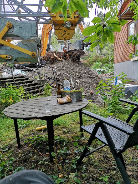 Uppgrävd trädgård med en grävmaskin, uppryckta trädrötter och ett bord med stolar under körsbärsblad i förgrunden.