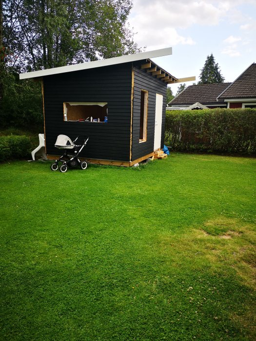 Svartmålat trädgårdsskjul under konstruktion med barnvagn framför på gräsmatta.